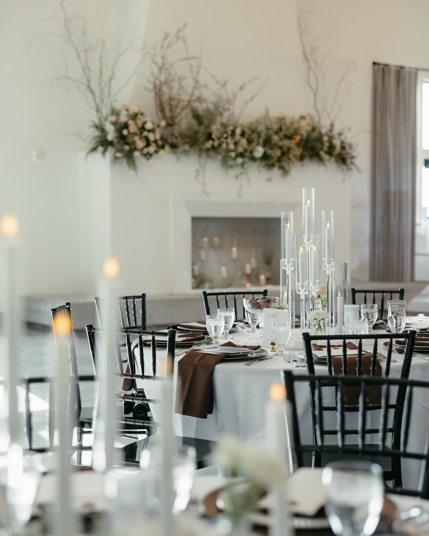 Elegant wedding reception table with black chiavari chairs, tall glass candelabras, and greenery garland above white fireplace mantel