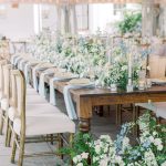 Outdoor garden wedding reception with long farm tables, greenery arrangements, and white tent