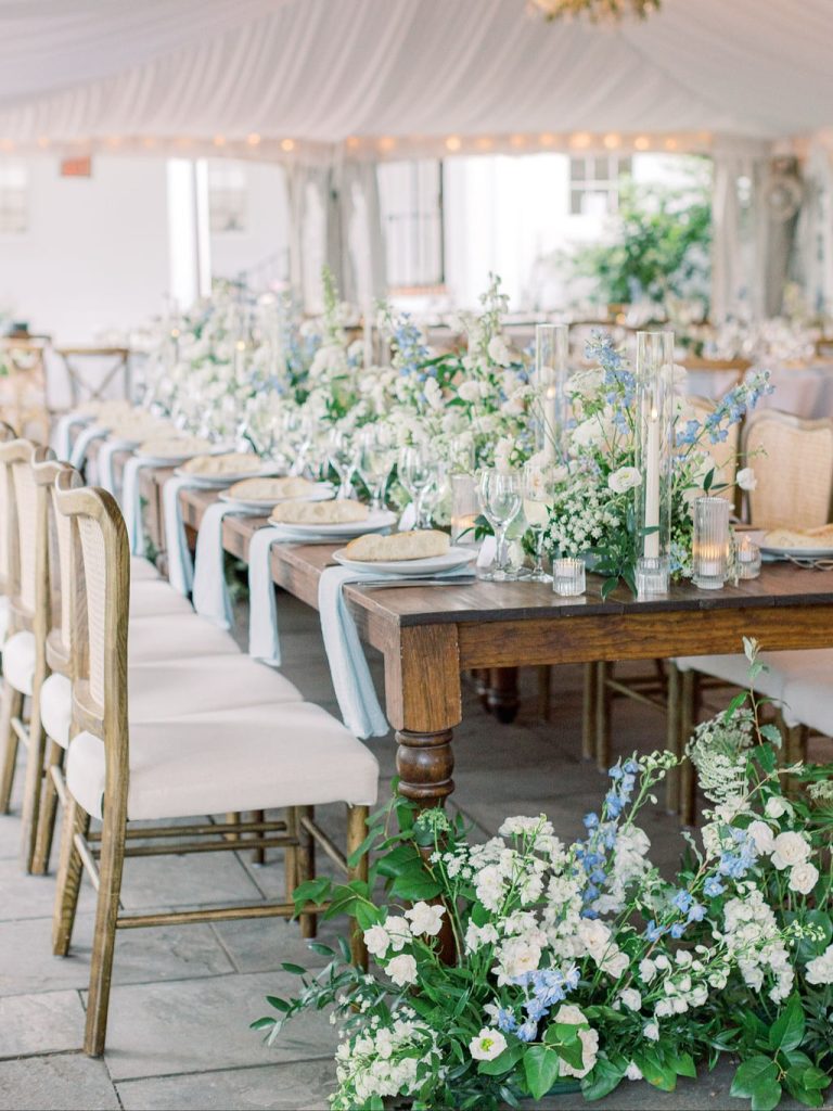 Outdoor garden wedding reception with long farm tables, greenery arrangements, and white tent