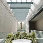 Modern atrium ceremony space with skylight panels, white circular platform surrounded by lush white and green floral arrangements