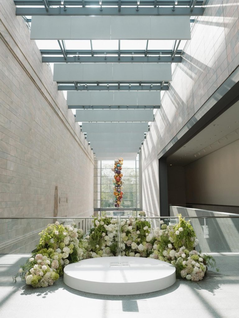 Modern atrium ceremony space with skylight panels, white circular platform surrounded by lush white and green floral arrangements