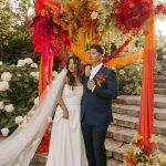 Newlyweds posing under elaborate floral arch with vibrant warm-toned flowers in garden ceremony space
