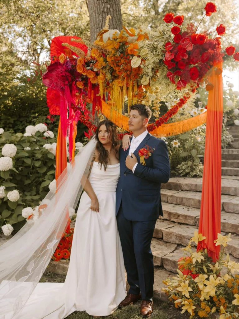Newlyweds posing under elaborate floral arch with vibrant warm-toned flowers in garden ceremony space