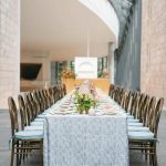 Long reception table with blue textured tablecloth, pink place settings, and floral centerpieces in modern venue with archway