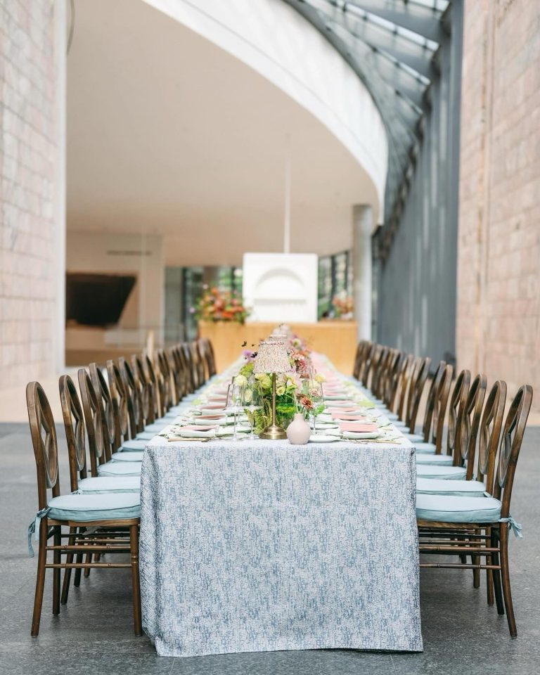 Long reception table with blue textured tablecloth, pink place settings, and floral centerpieces in modern venue with archway