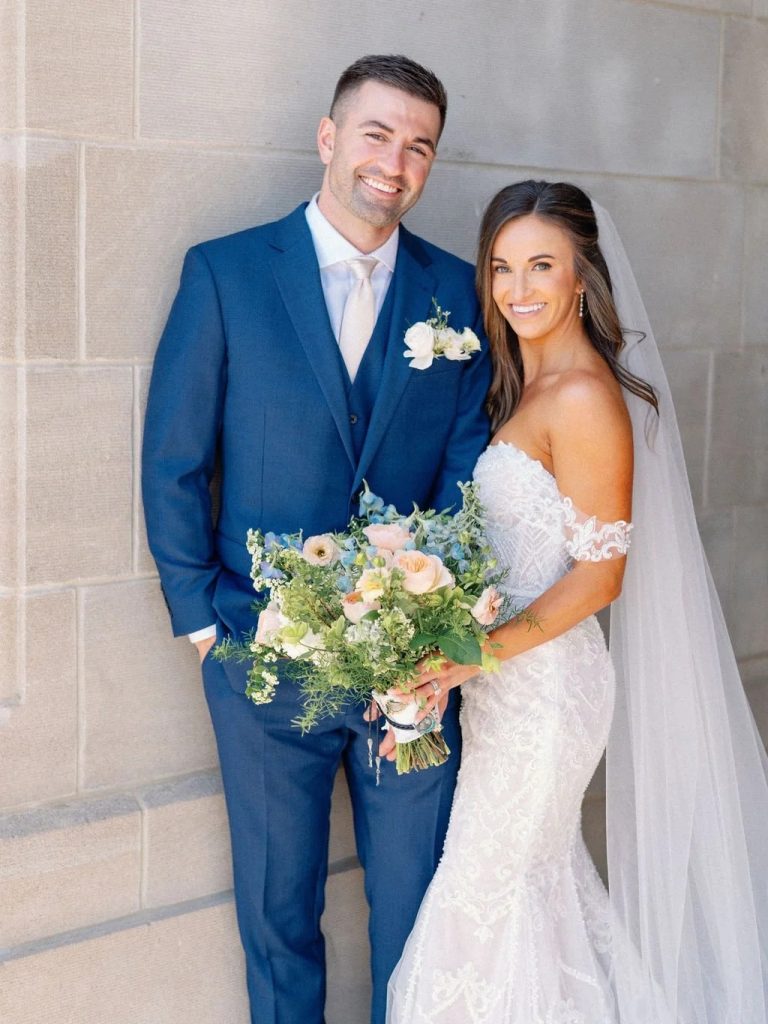 Newlyweds smiling together, groom in navy suit and bride in lace gown with pastel bridal bouquet