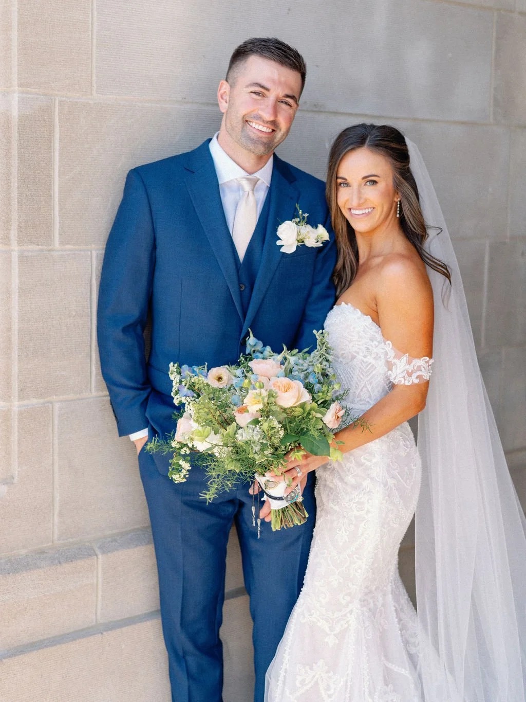 Newlyweds smiling together, groom in navy suit and bride in lace gown with pastel bridal bouquet