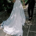 Bride in white lace wedding gown with train holds groom's hand on stone patio framed by ornamental iron gate and greenery