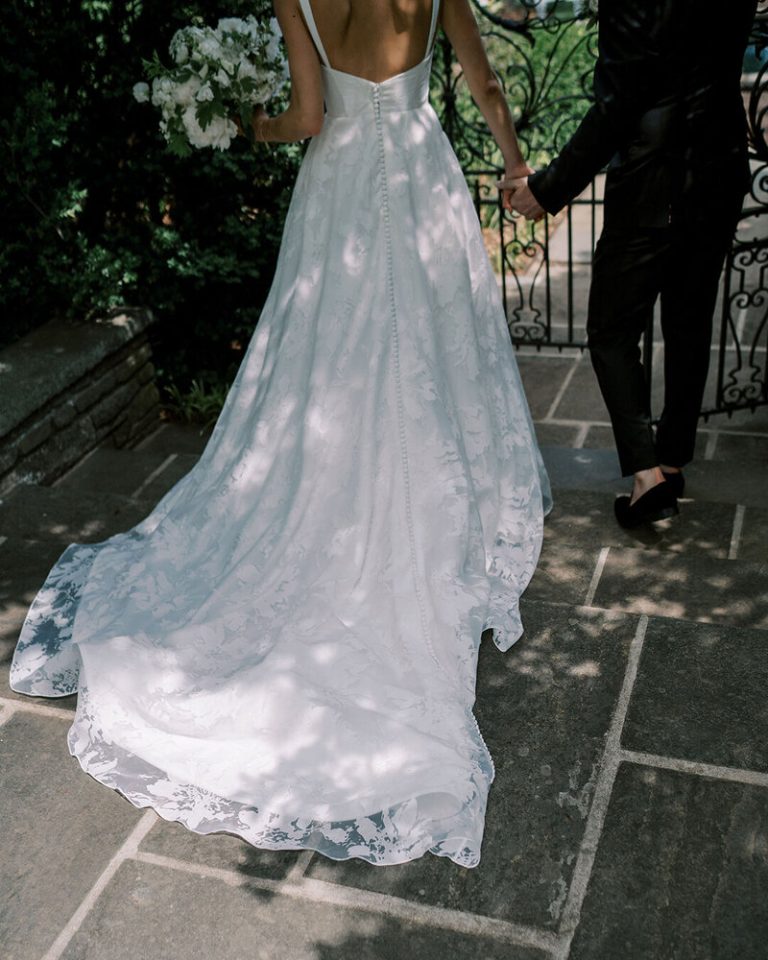 Bride in white lace wedding gown with train holds groom's hand on stone patio framed by ornamental iron gate and greenery