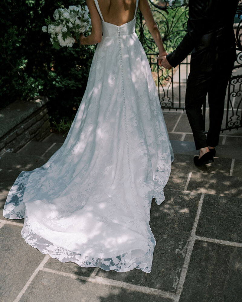 Bride in white lace wedding gown with train holds groom's hand on stone patio framed by ornamental iron gate and greenery