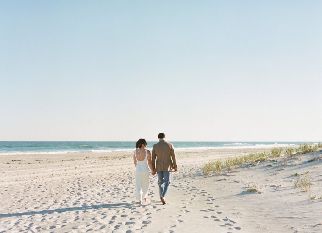 Newlywed couple holding hands walking along sandy beach toward ocean at sunset