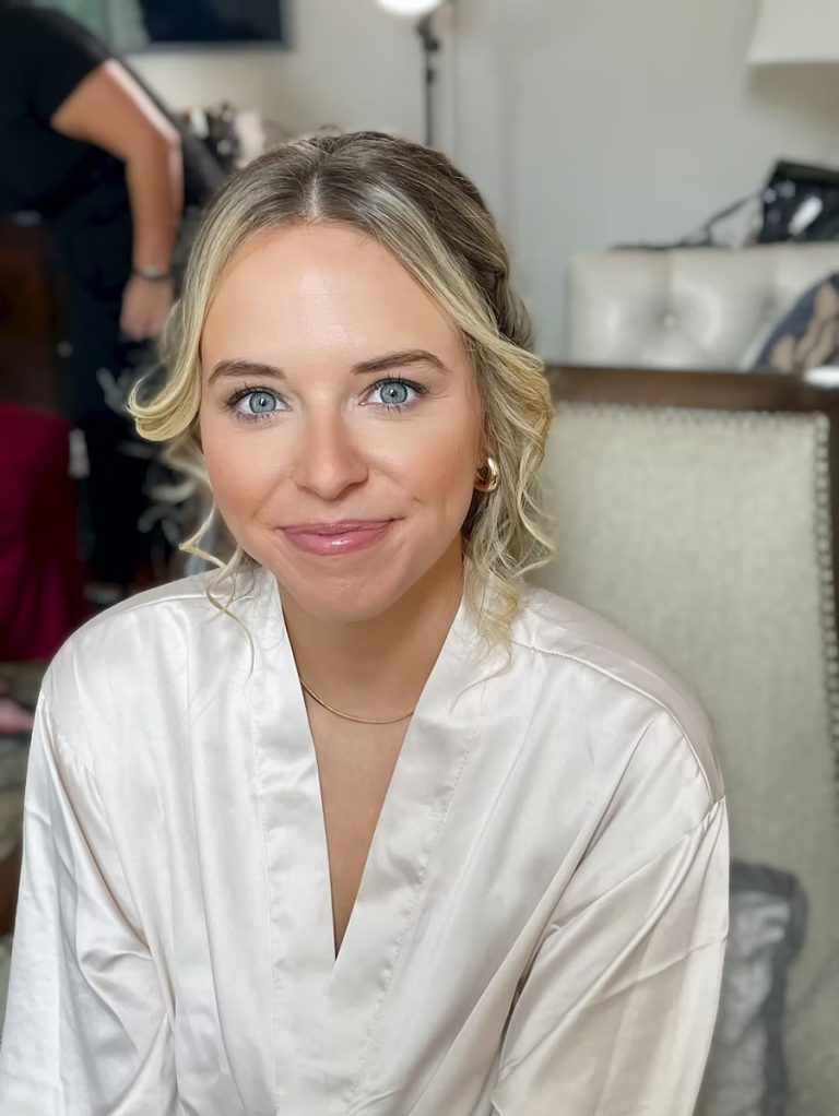 Blonde bride in white satin robe with soft curled updo during wedding preparation
