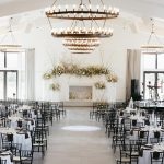 Elegant white wedding reception hall with tiered circular chandeliers, black chiavari chairs, and floral-adorned fireplace mantel