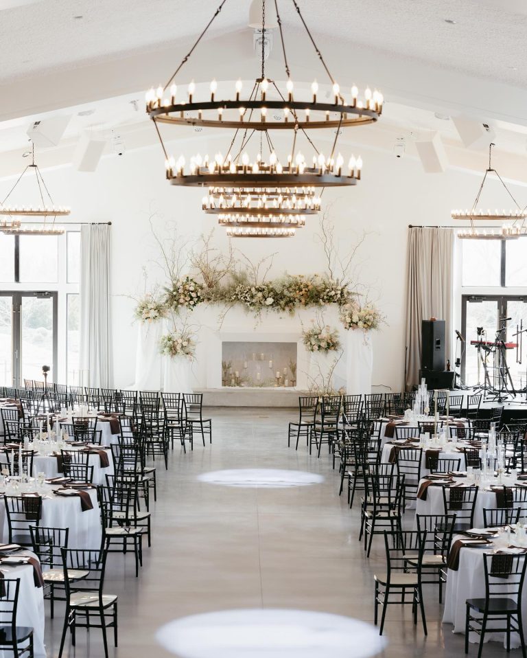 Elegant white wedding reception hall with tiered circular chandeliers, black chiavari chairs, and floral-adorned fireplace mantel