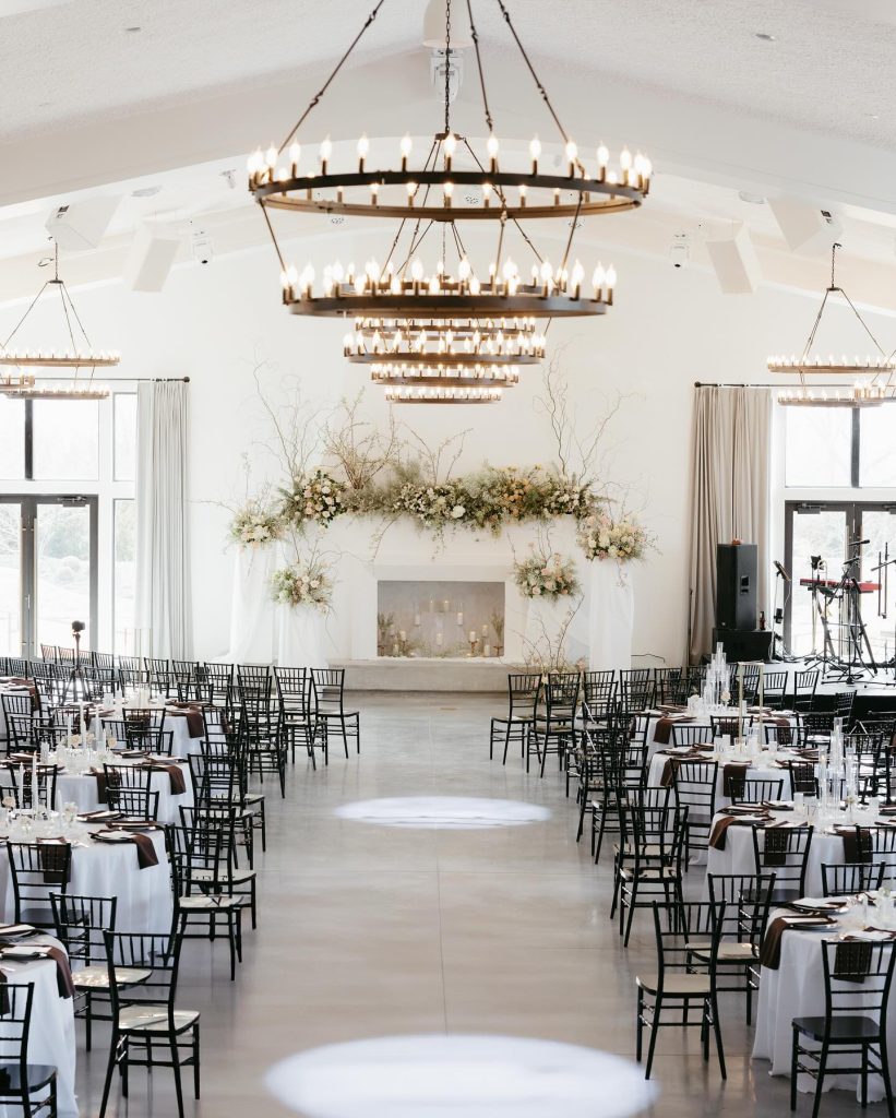 Elegant white wedding reception hall with tiered circular chandeliers, black chiavari chairs, and floral-adorned fireplace mantel