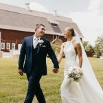 Bride and groom walk hand-in-hand across lawn with white barn in background