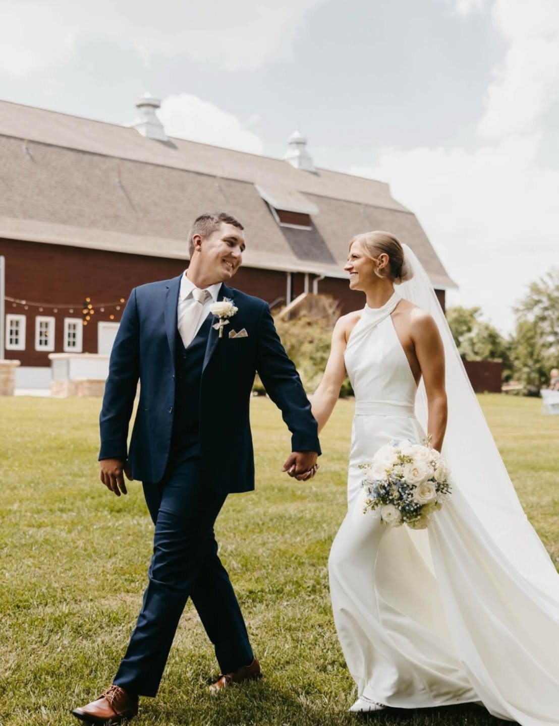 Bride and groom walk hand-in-hand across lawn with white barn in background