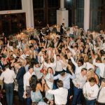 Crowded dance floor filled with wedding guests dancing with hands raised under chandelier and string lights