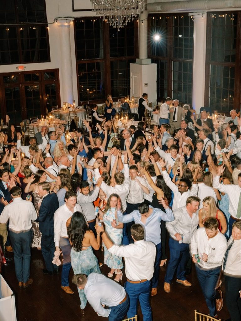 Crowded dance floor filled with wedding guests dancing with hands raised under chandelier and string lights