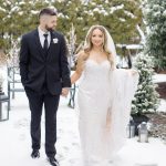 Smiling bride in beaded gown with groom in black suit strolling through snowy outdoor venue