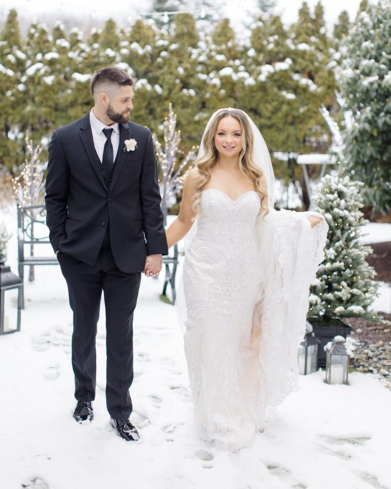 Smiling bride in beaded gown with groom in black suit strolling through snowy outdoor venue
