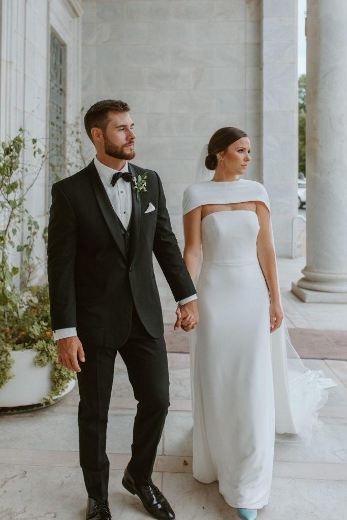 Bride in off-shoulder gown and groom in black tuxedo walk hand-in-hand between white columns