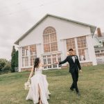 Bride and groom walking hand in hand on lawn in front of white chapel with arched windows