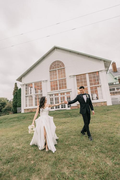 Bride and groom walking hand in hand on lawn in front of white chapel with arched windows