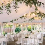 Elegant white wedding tent reception with crystal chandeliers, greenery garlands, and gold chiavari chairs