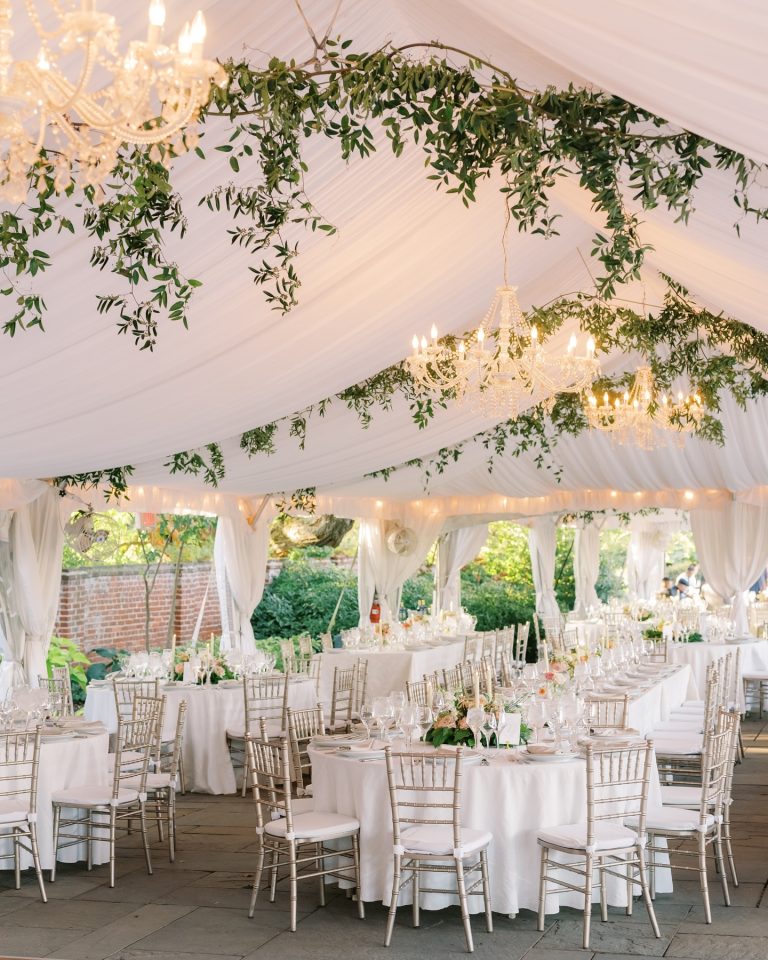 Elegant white wedding tent reception with crystal chandeliers, greenery garlands, and gold chiavari chairs