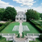 Aerial view of elegant colonial mansion with white folding chairs arranged for outdoor ceremony on manicured lawn with formal gardens