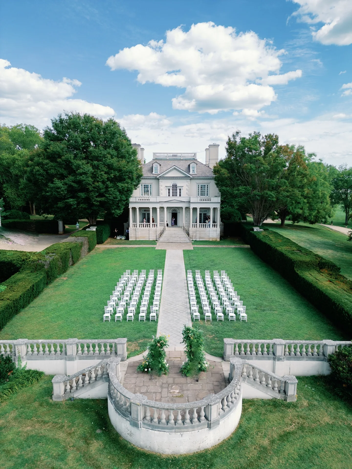 Aerial view of elegant colonial mansion with white folding chairs arranged for outdoor ceremony on manicured lawn with formal gardens