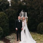 Newlywed couple embracing in front of white decorative gazebo surrounded by trees