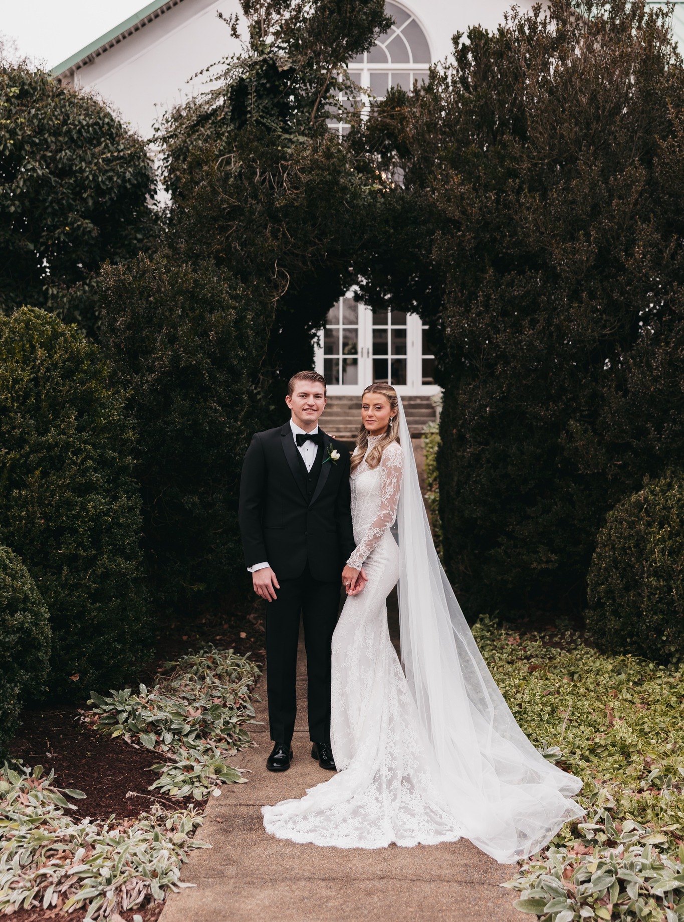 Newlywed couple embracing in front of white decorative gazebo surrounded by trees