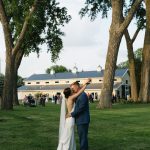 Bride and groom embrace on lawn under mature trees with barn-style Nebraska wedding venue in background