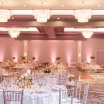 Elegant ballroom reception space with coffered ceiling, statement chandeliers, round tables, and rose gold accent lighting