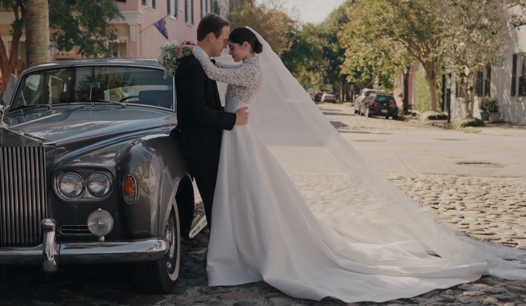 Bride and groom embracing beside vintage luxury car on tree-lined cobblestone street