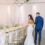 Bride and groom walk beside elegantly set reception table with gold chiavari chairs and hanging greenery with string lights