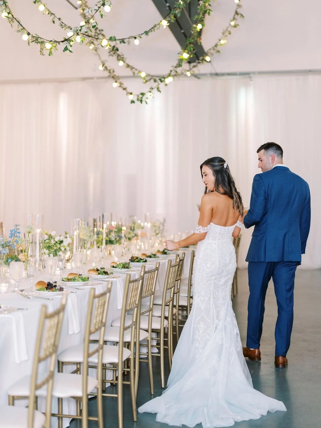 Bride and groom walk beside elegantly set reception table with gold chiavari chairs and hanging greenery with string lights