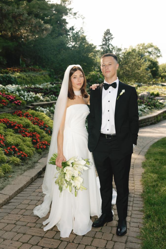 Bride in strapless gown with veil and groom in black tuxedo standing on brick pathway surrounded by terraced gardens