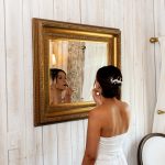 Bride in strapless white gown applying lipstick while looking in ornate gold mirror against white wooden wall