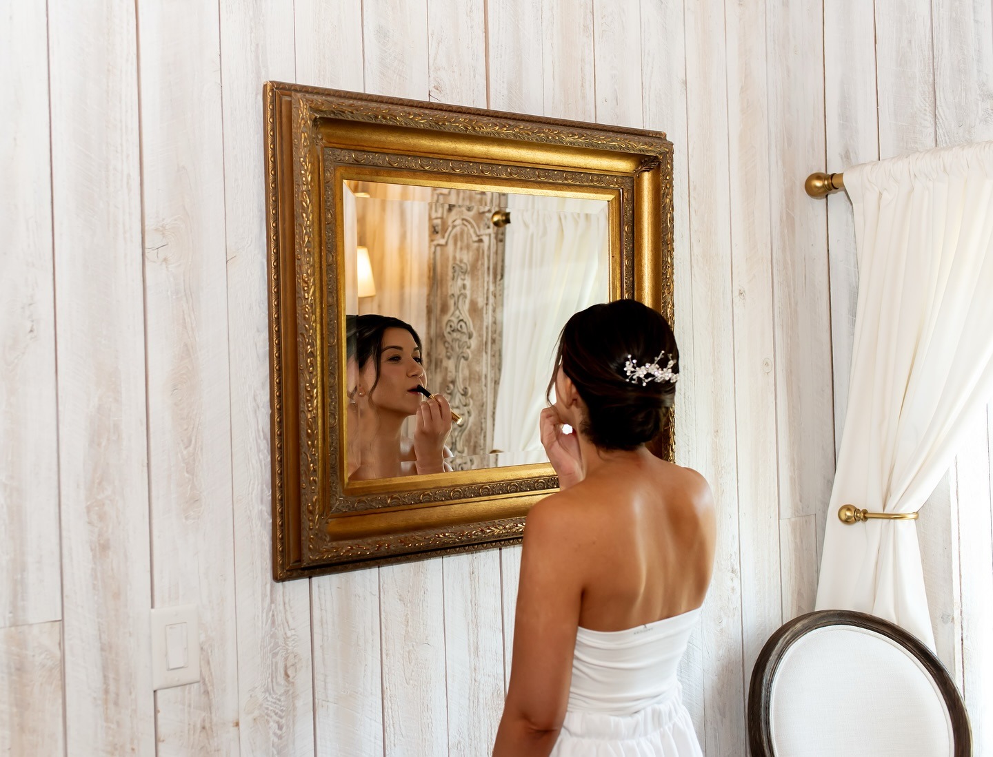 Bride in strapless white gown applying lipstick while looking in ornate gold mirror against white wooden wall