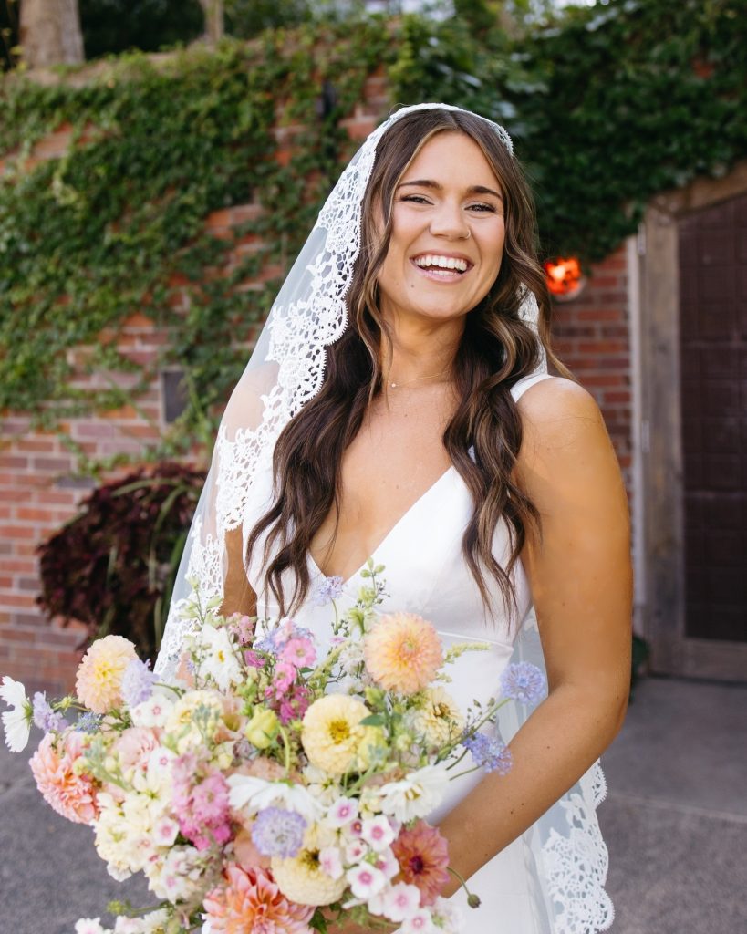 Joyful bride in lace-edged veil holds abundant pastel bouquet in garden setting