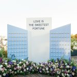 Blue and white escort card display with fortune cookie favors and "Love is the Sweetest Fortune" sign adorned with vibrant floral arrangement