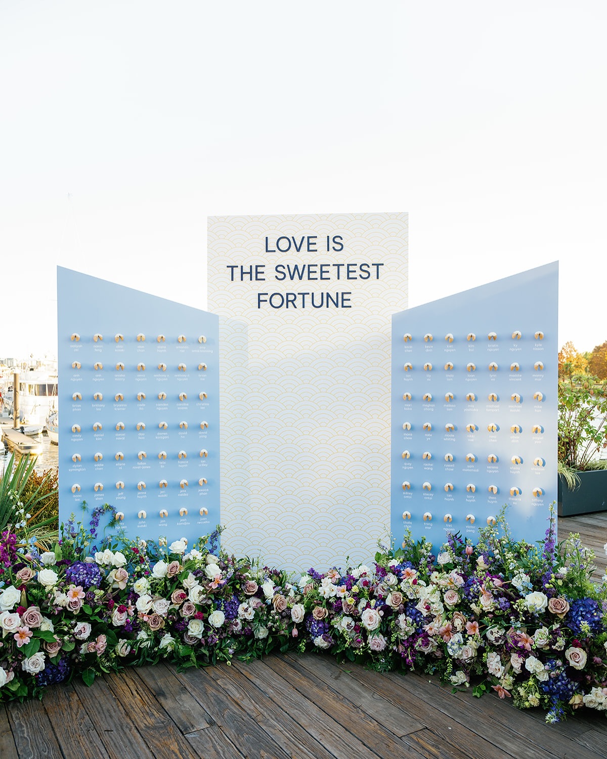 Blue and white escort card display with fortune cookie favors and "Love is the Sweetest Fortune" sign adorned with vibrant floral arrangement
