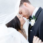 Bride and groom sharing an intimate moment under flowing veil, groom wearing black suit with white boutonniere