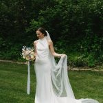 Bride in halter-neck gown holding pastel bouquet on manicured lawn with forest backdrop