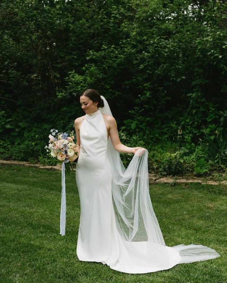 Bride in halter-neck gown holding pastel bouquet on manicured lawn with forest backdrop