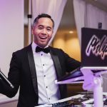 Wedding DJ in tuxedo with microphone standing behind illuminated equipment booth