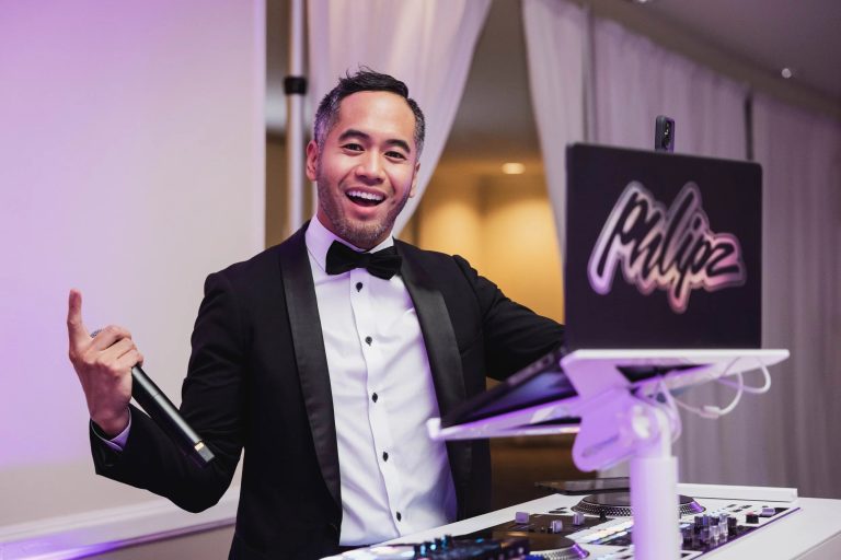 Wedding DJ in tuxedo with microphone standing behind illuminated equipment booth